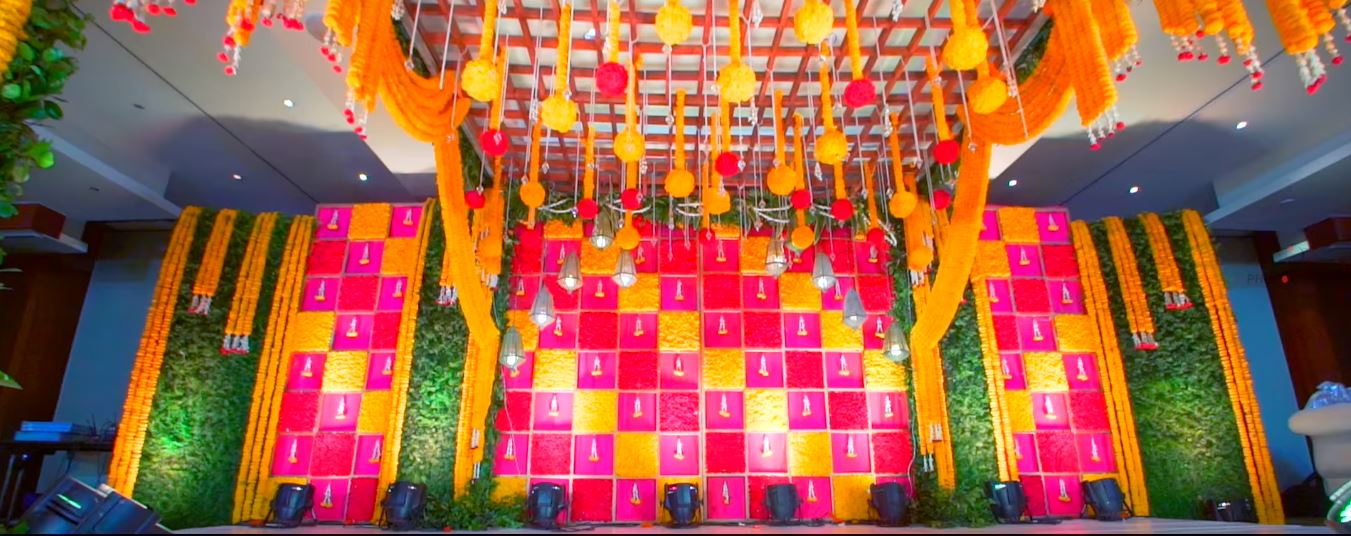 Hanging marigold balls and strings below a wooden lattice ceiling, above a yellow and pink flower-panel backdrop with green foliage walls