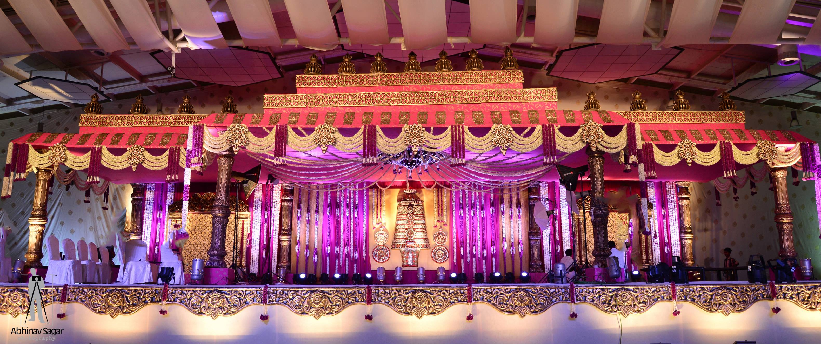 Panoramic indoor wedding stage with tall carved gold pillars, pink and gold canopies, jasmine strands and a central jewelled backdrop under a draped ceiling