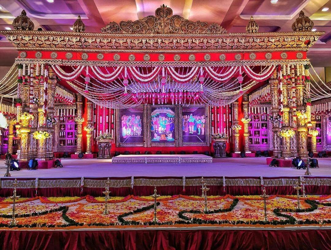 Grand red and gold carved mandap with painted mythological panels, jasmine swags, brass lamps and a flower-petal rangoli in the foreground