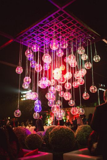 Dozens of glass baubles with candles hanging on strings from a wooden lattice, lit in pink and purple above topiary balls
