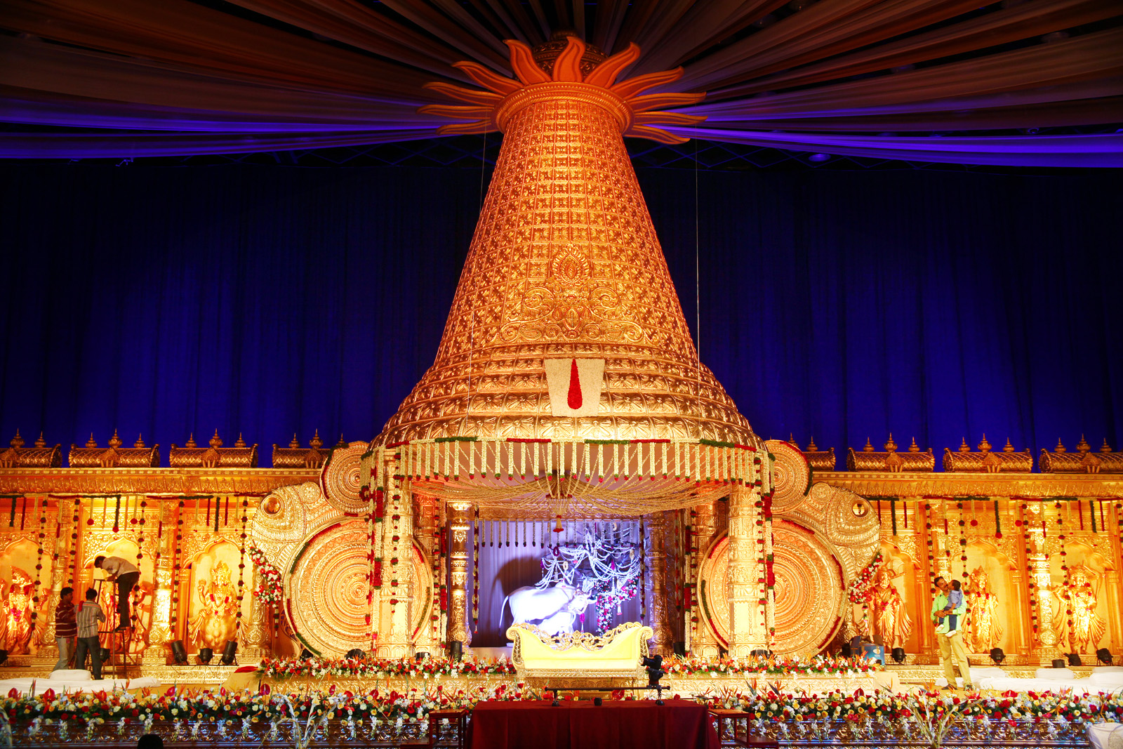 Towering golden conical mandap shaped like a temple tower, lit warm against deep blue drapes, with a white and gold love-seat beneath and flowers along the stage edge