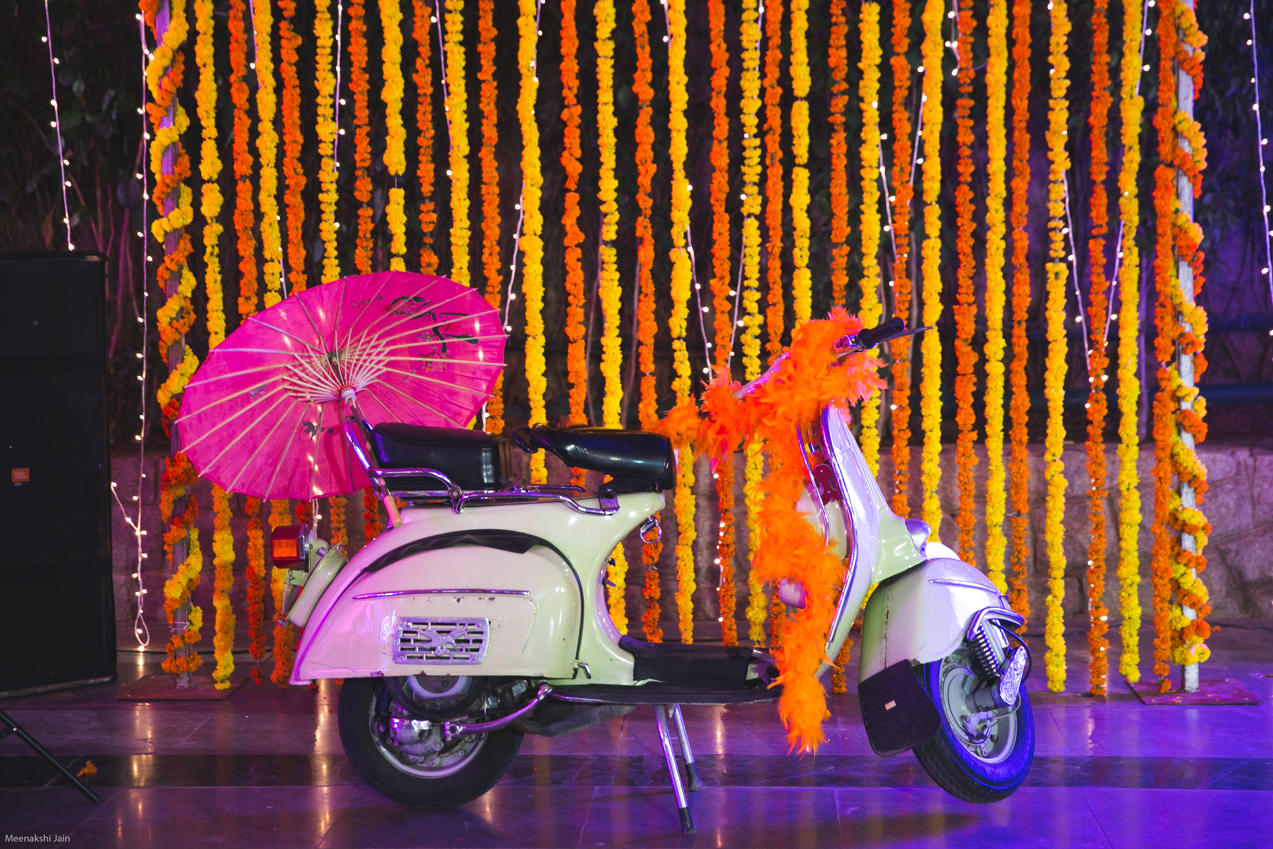 Cream vintage scooter with a pink paper parasol and orange feather boa in front of a wall of hanging marigold strings and fairy lights