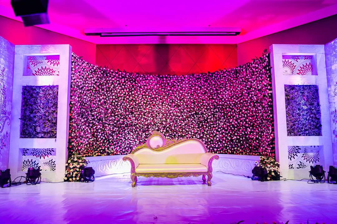 Curved wall of pink and white roses behind a white and gold sofa on a white stage, lit in magenta, flanked by white panels with cut-out patterns