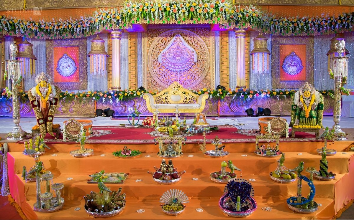 Traditional South Indian wedding stage with carved gold panels, a white and gold love-seat, silver lamps and ritual trays arranged on orange-draped steps