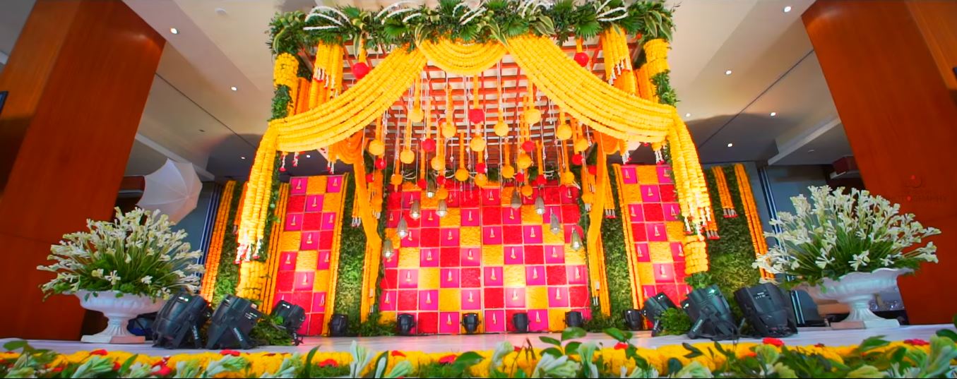 Daytime celebration stage with a chequerboard backdrop of yellow and pink flower panels, marigold swags on a lattice canopy, green foliage walls and white urns of flowers