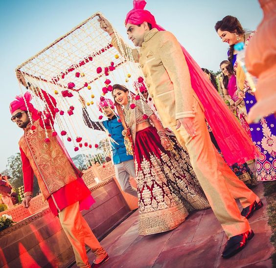A bride in a maroon lehenga walks beneath a canopy of white jasmine strings and pink roses held by four men in cream and gold, outdoors in daylight
