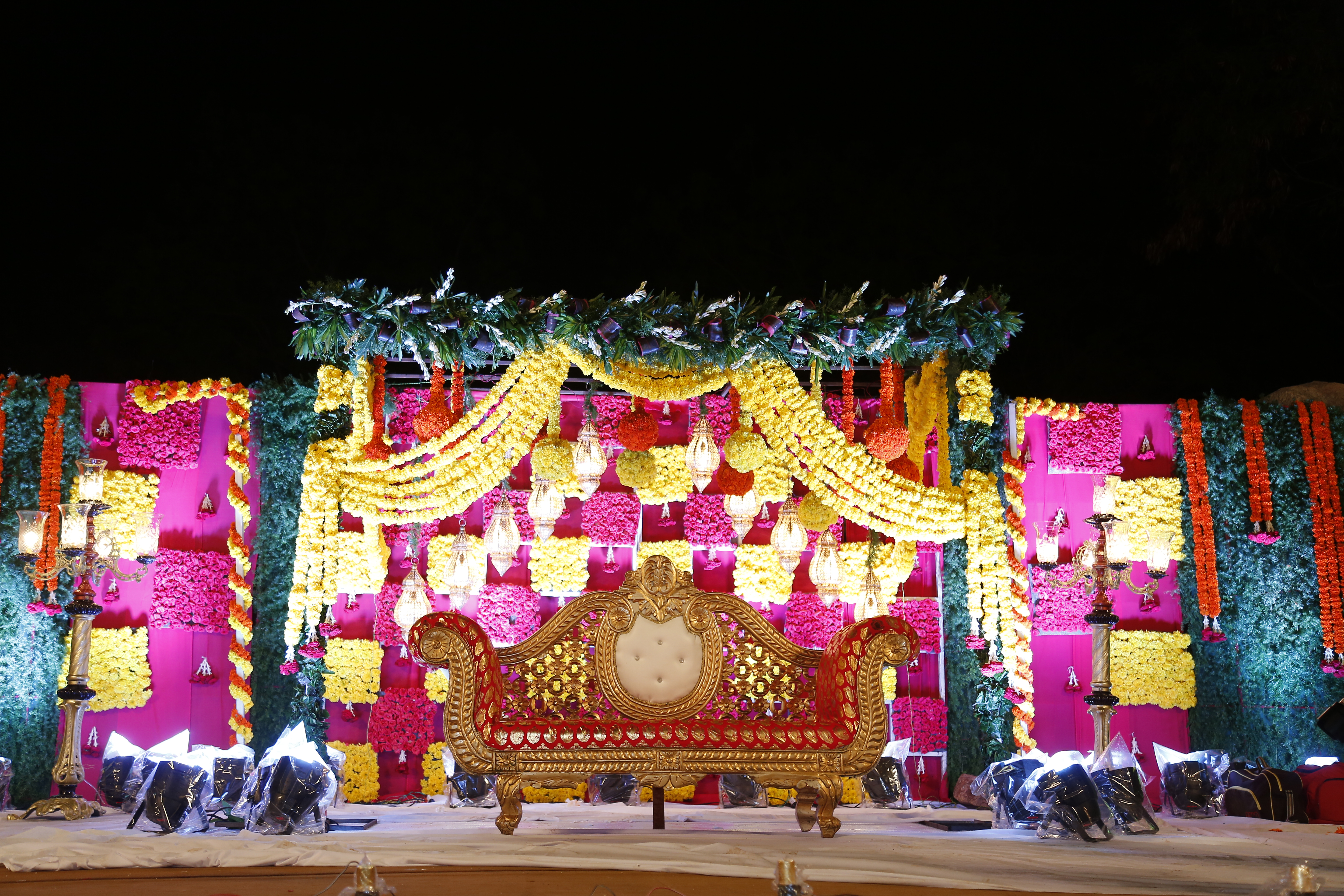 Outdoor evening stage dressed in pink and yellow floral panels with hanging lanterns, marigold swags and a carved gold love-seat under a canopy of foliage