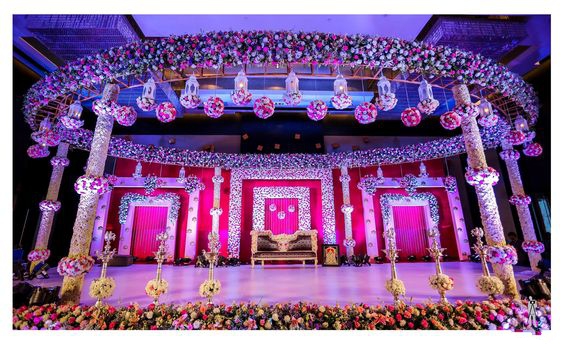 Circular floral mandap with pink and white blooms on the canopy ring and pillars, hanging lanterns and a gold love-seat, framed in white